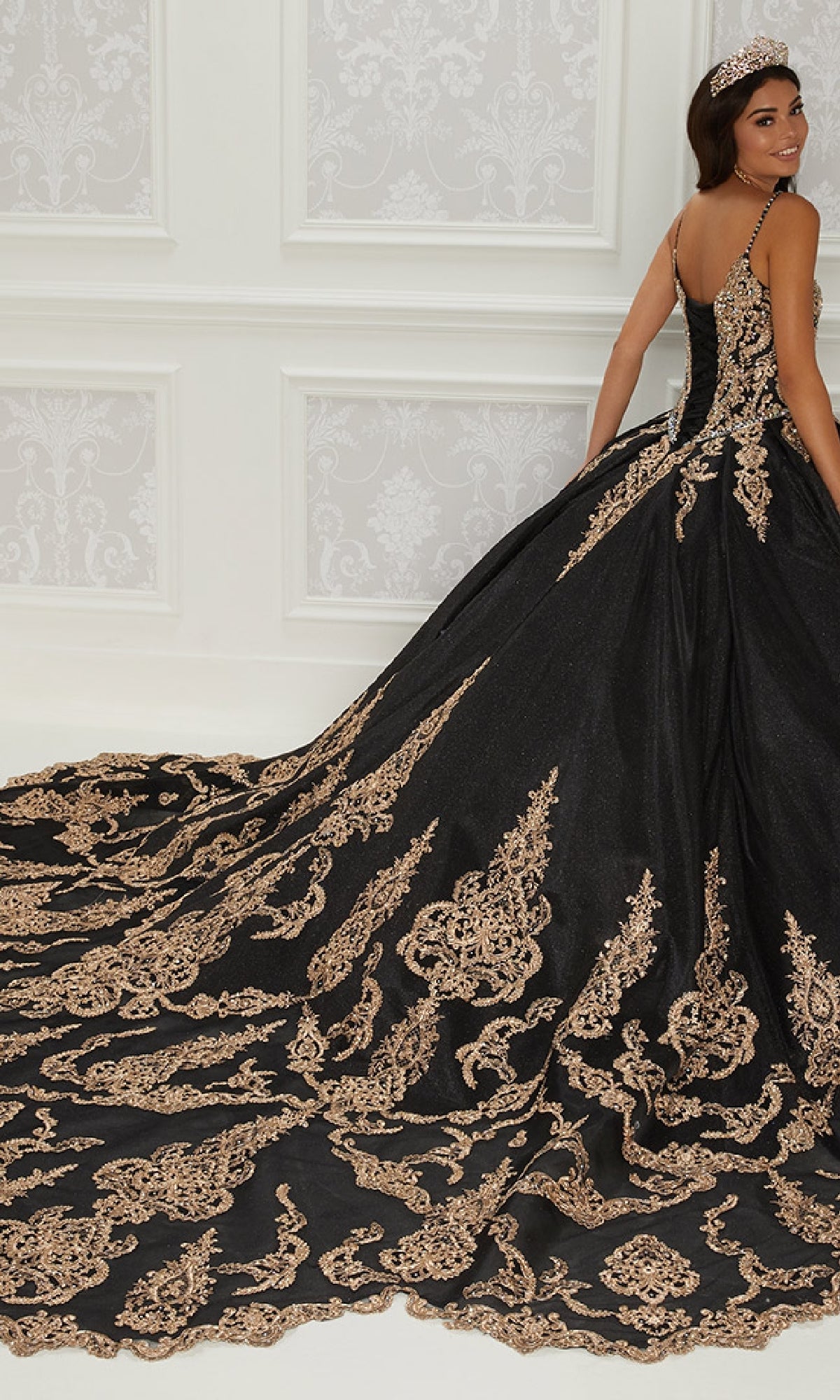 A woman in the Quinceanera Dress PR22141 By Princesa—black with gold embroidery, corset back, and long train—stands in an ornate room, facing slightly away from the camera.
